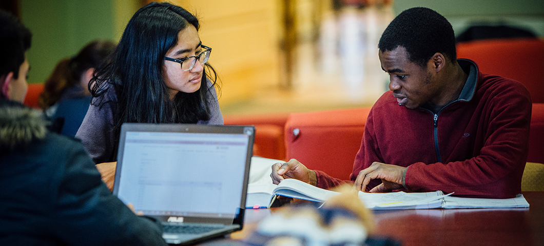 Two students work around a laptop in a building lobby.