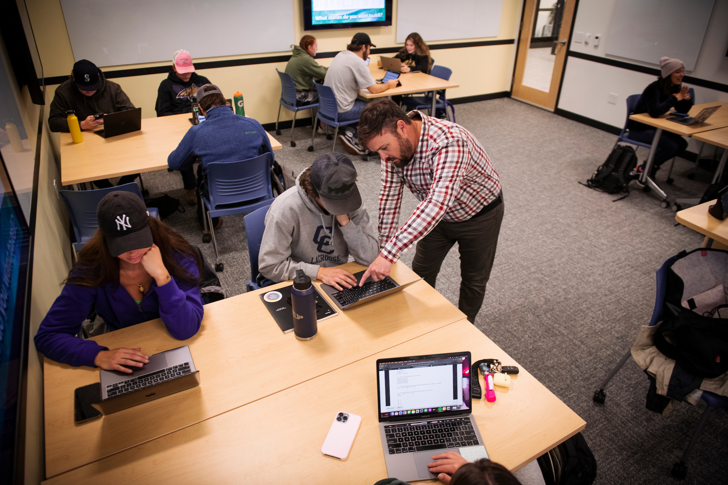 Instructor leans over a table to point at a student’s laptop while several college students work independently or in small groups on laptops in a classroom.