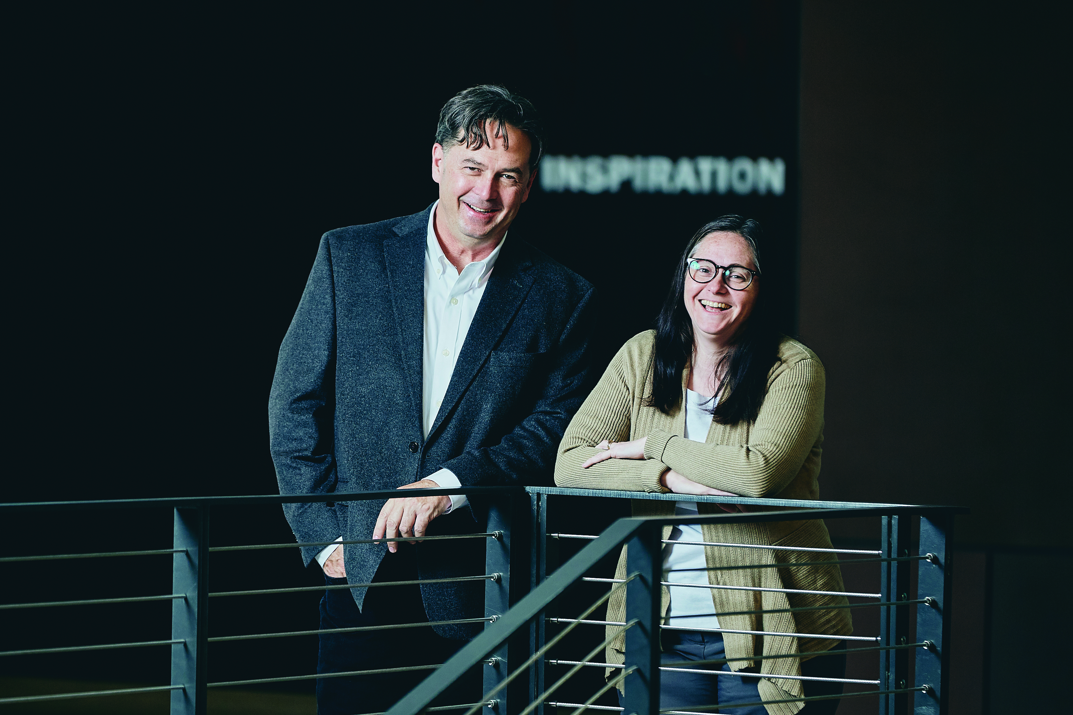 A person with short dark hair and a dark suit coat next to a person wearing glasses and a light brown cardigan