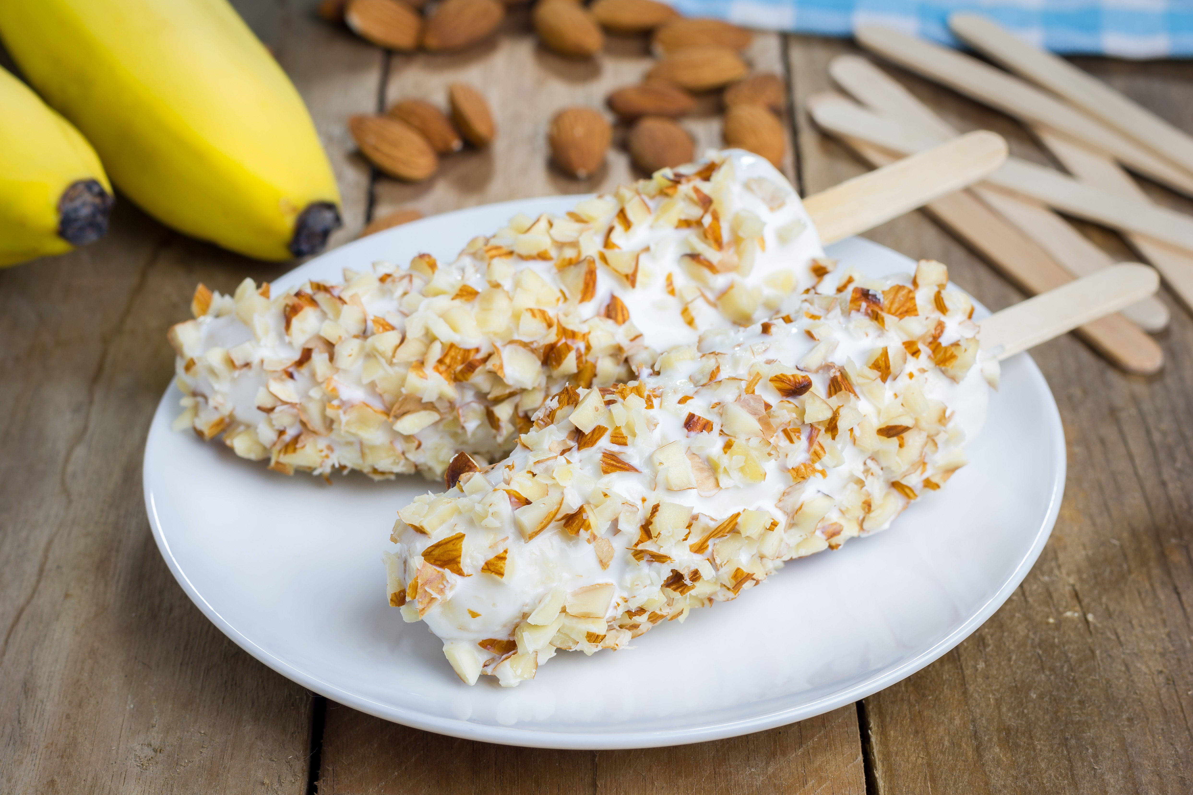 A white plate with two bananas on popsicle sticks, covered in yogurt and chopped nuts.