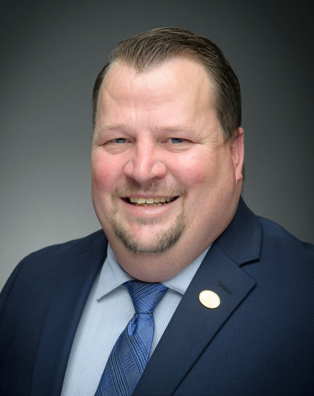 Cody Stone in a navy suit jacket with a light blue button up and blue tie on. There is a circular, gold pin attached to his left suit lapel.