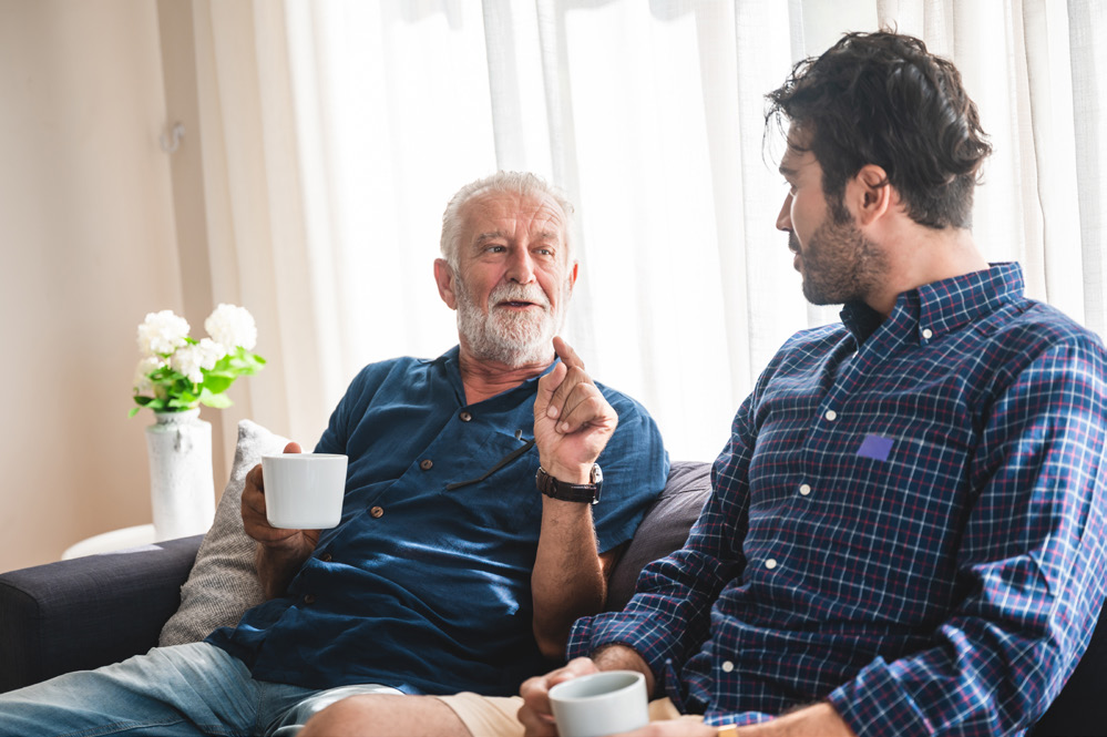 Two men sit drinking coffee, one is aged with white hair and a white beard and the other is younger.