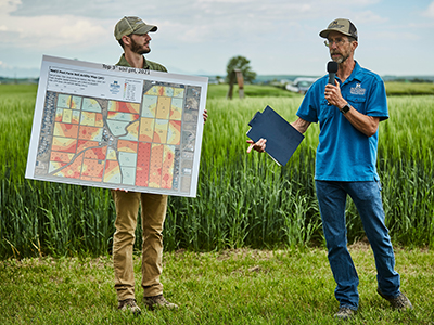 MSU Extension: A Partner in Meeting Local Needs Two people stand in a grassy field during an outdoor agricultural event. One person holds a large map showing color-coded field plots, while the other speaks into a microphone and holds a folder. Tall green crops and a cloudy sky are visible in the background.