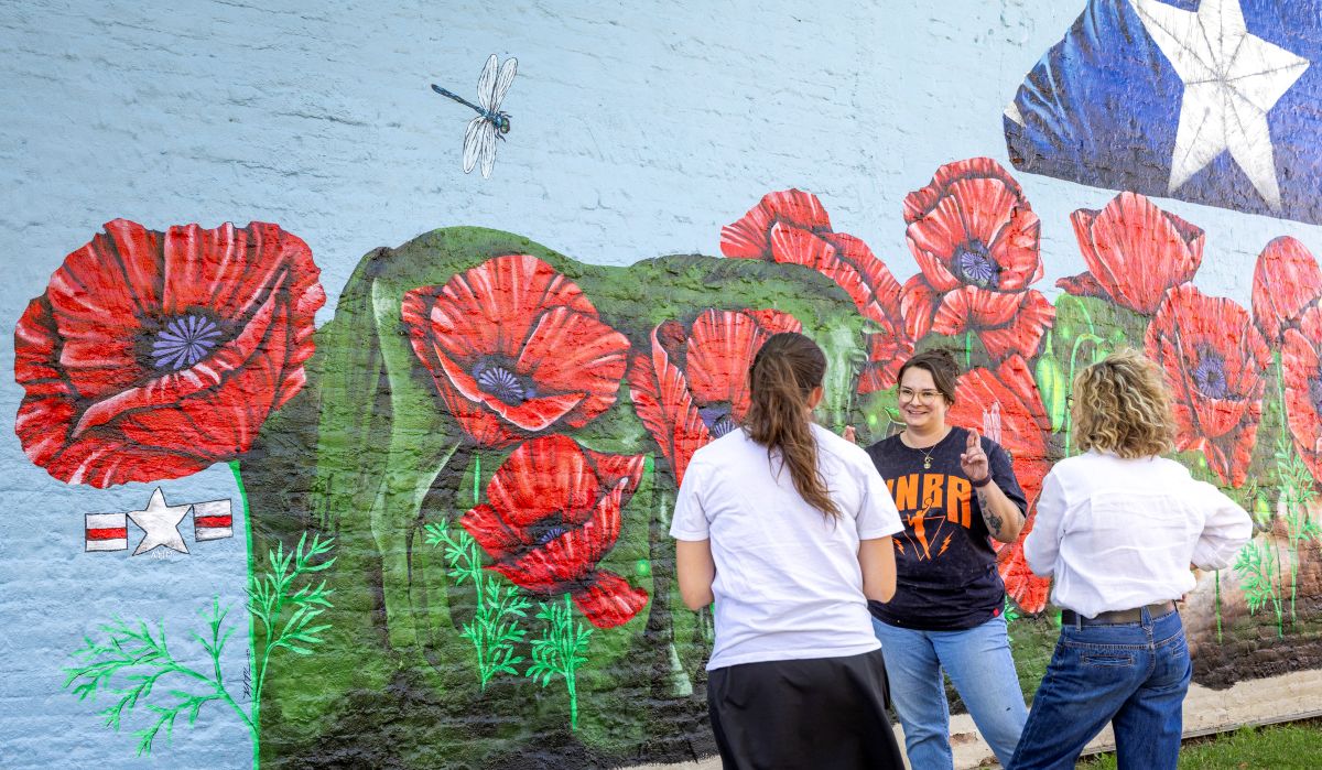 Three people standing in front of the Veteran's Park mural in Miles City. In the photo, the mural features you can see inlcude large red poppy flowers, green stems, a dragonfly, and the white star on blue background of a tri-corner folded burial flag.