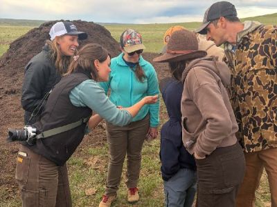 Winnett ACES Participants gathered to examine a soil sample while standing near compost piles in open grassland.