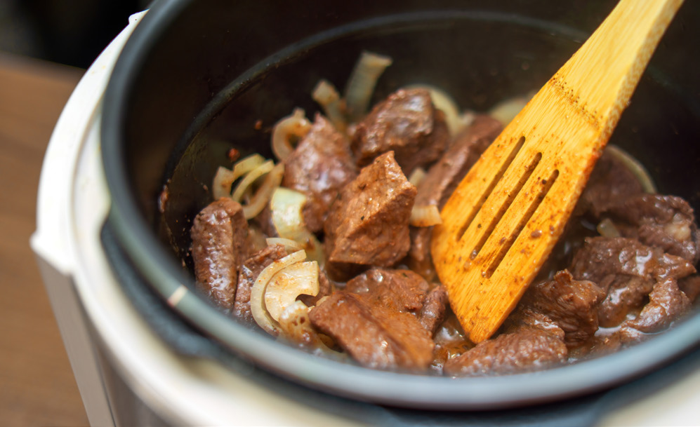 A slotted wooden spatula rests inside a pressure cooker full of seasoned meat and vegetables
