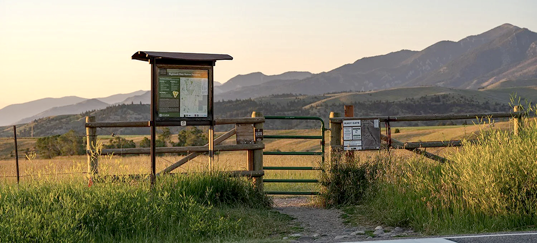 A trailhead with cattle gate on the Highland Glen property in the summer is flanked by tall grasses and trail maps and information. The Bridger Mountains feature prominantly in the background.