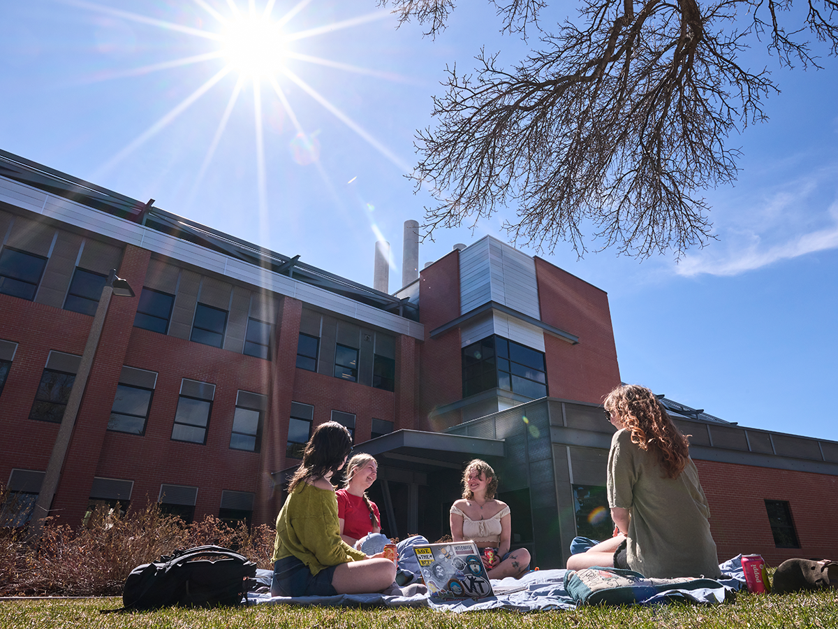 A small group of students sitting in grass with laptops around them. There is brick buildings and a bright sunny sky in the background