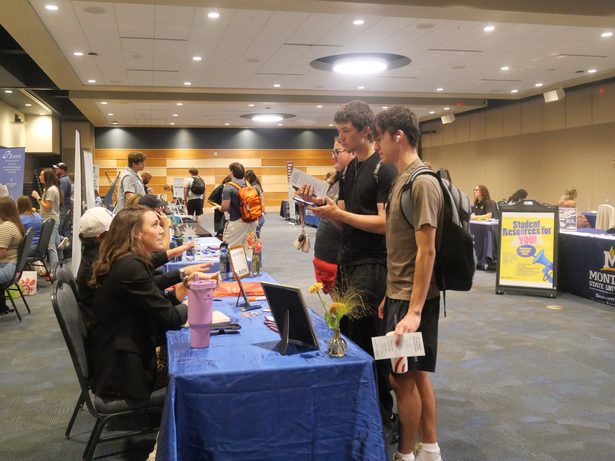 Two students wearing backpacks talking to two people sitting at a table with materials on it