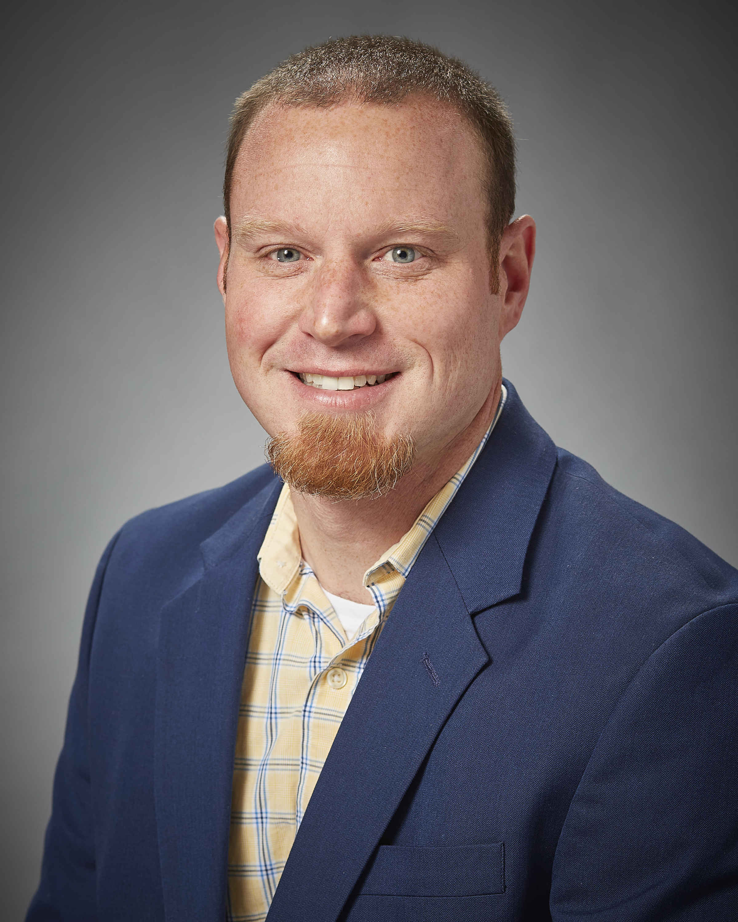 A portrait of a person with short hair and a medium-short beard, wearing a yellow button up shirt with a blue suit jacket