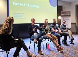 A group of five panelists sitting on a stage in front of a screen. One is holding a microphone.
