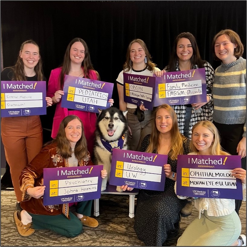 Eight Montana WWAMI students celebrate Residency Match Day 2026 by holding signs indicating where they matched. A husky dog sits in the center of the group. 